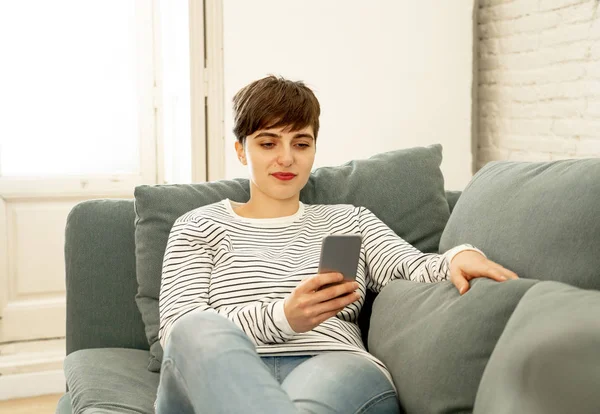 快乐的有吸引力的短发年轻女子在手机聊天 并使用社交媒体应用程序坐在家里的沙发上约会在线约会 使用新技术和社交网络的概念 — 图库照片