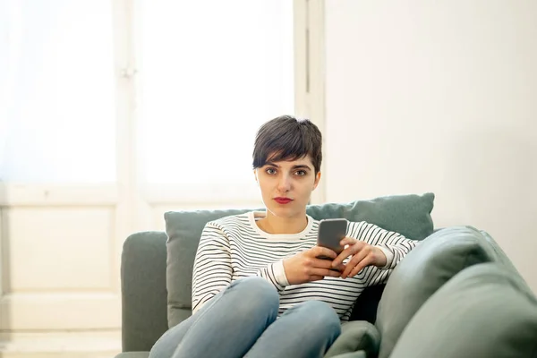 Happy attractive short haired young woman on mobile smart phone chatting and using social media apps sitting on couch at home in dating on line,using new technology and social network concept.