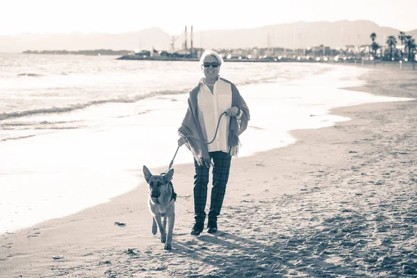 Beautiful retired older woman and pet german shepard dog walking along the shore sea ocean on beach in Companionship Benefits of animals Keeping active Retirement lifestyle and Dog friendly tourism.