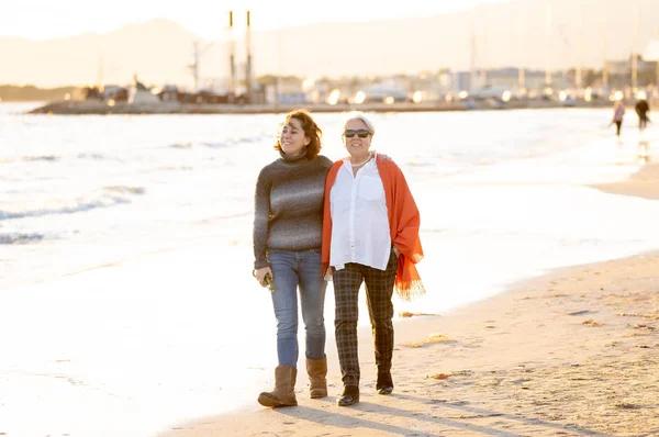 愉快的高级母亲和成年女儿一起度过的画像手牵手笑和走在海滩上日落光在快乐的家庭时刻世代退休和人的概念 — 图库照片
