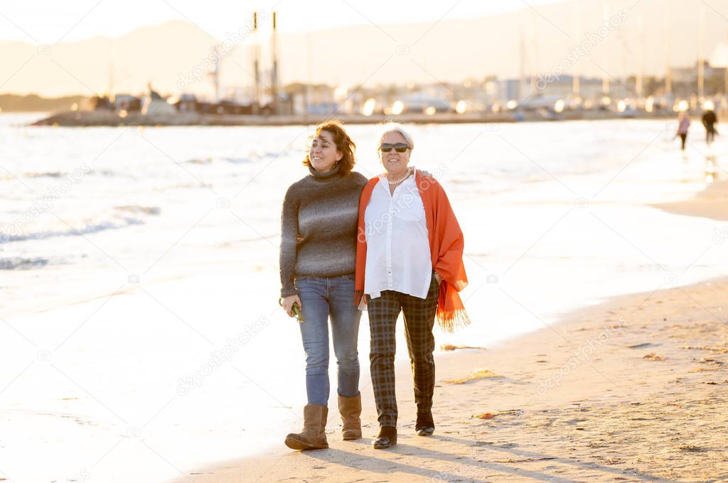 Portrait of happy senior mother and adult daughter spending time together holding hands laughing and walking on beach at sunset light in Happy family moments Generations Retirement and People concept.