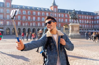 Gülümseyen genç beyaz turistik öğrenci adam mutlu ve heyecanlı bir selfie akıllı cep telefonu ile Plaza Mayor, Madrid, İspanya alarak eğleniyor. Seyahat, tatil, tatil Europe kavramı çevresinde.