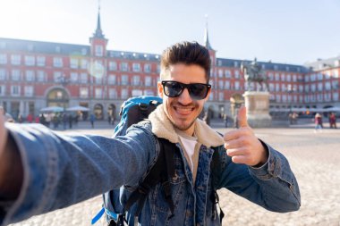 Çekici genç turist öğrenci adam mutlu ve heyecanlı bir komik surat selfie akıllı telefon ile Plaza Mayor, Madrid, İspanya alarak eğlenmeye sırt çantası. Tatil ve seyahat etmek çevrede dünya kavramı.
