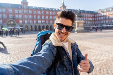 Çekici genç turist öğrenci adam mutlu ve heyecanlı bir komik surat selfie akıllı telefon ile Plaza Mayor, Madrid, İspanya alarak eğlenmeye sırt çantası. Tatil ve seyahat etmek çevrede dünya kavramı.