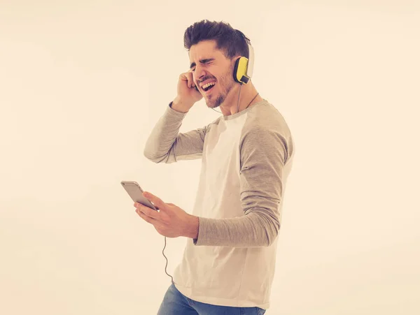 Portrait of happy young man listening to music in headphones changing songs and tracks on smartphone and singing and dancing. Isolated white background. In positive emotions, youth happiness concept.