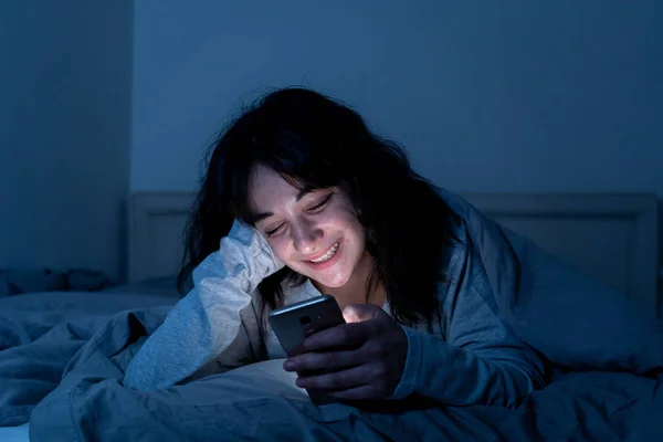 Привлекательная латинская женщина, пристрастившаяся к мобильному телефону и интернету поздно ночью в постели, выглядит бессонной — стоковое фото
