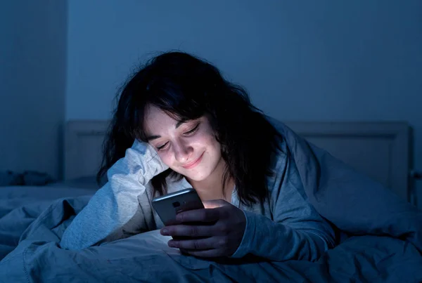 Привлекательная латинская женщина, пристрастившаяся к мобильному телефону и интернету поздно ночью в постели, выглядит бессонной — стоковое фото