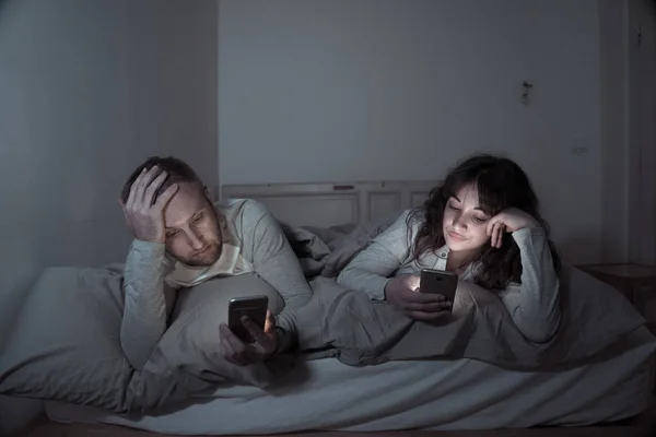 Life style portrait of young bored couple in bed at night on smart phones obsessed with games, social media, apps ignoring each other. Relationship communication problems and phone addiction concept.
