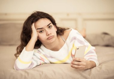 Closeup of young beautiful bored girl on bed using mobile phone. Tired checking smartphone in melancholic mood. Communication internet and technologies addiction and overuse, Cell phone dependency. clipart