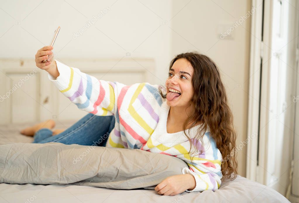 Excited trendy young teenager woman taking a selfie on smart mobile phone on bed at home. Happy talking and posting online in social media apps. In new technology, connection and Internet concept.