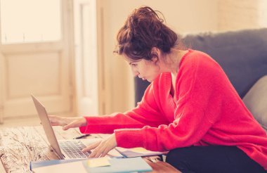 Akıllı bir kırmızı kazak çalışma giyen ve evde kanepede oturan bir dizüstü bilgisayar üzerinde okuyan bir düşünceli Casual güzel kadın yakın. Evde çalışma ve Internet kavramı üzerinde sörf.