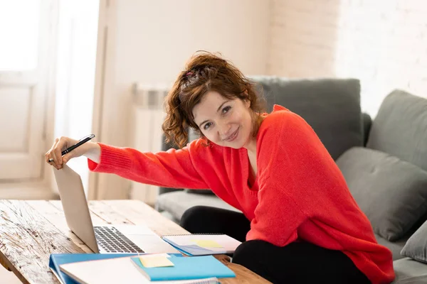 一个开朗的休闲美丽的女人穿着一个明亮的红色套头衫工作和学习的笔记本电脑坐在沙发上在家里 在家里工作和上网的概念 — 图库照片