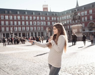 Güzel ve neşeli genç kadını mutlu ve heyecanlı başparmak yukarıya jest yapma akıllı Plaza Mayor Madrid alarak selfie. Avrupa, İspanya ve sosyal medya turizm öğrenci yaşam, seyahat.