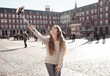 Güzel genç öğrenci kadın mutlu ve heyecanlı Plaza Mayor Madrid alarak selfie veya video mobil akıllı telefon selfie sopa ile. Bir öpücük kameraya üfleme. Avrupa İspanya'da turizm, seyahat.