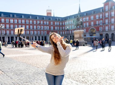 Güzel genç kadın mutlu ve heyecanlı Plaza Mayor Madrid İspanya alarak selfie veya video tarihi mimari zevk onun takipçileri için poz selfie sopa ile. Turizm ve seyahat Europe.