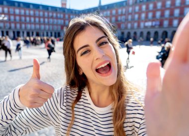 Beautiful young student tourist woman happy and excited in Plaza Mayor Madrid taking a selfie holding the mobile or tablet. Looking cheerful making thumbs up gesture. In tourism and travel in Europe. clipart