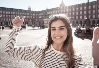 Beautiful young student tourist woman happy and excited in Plaza Mayor Madrid taking a selfie holding the mobile or tablet. Looking cheerful making thumbs up gesture. In tourism and travel in Europe. clipart
