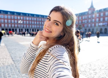 Beautiful student tourist woman happy and excited taking close up selfie in Plaza Mayor Madrid Looking cheerful and joyful having fun. In tourism, travel around europe and posting online adventures. clipart