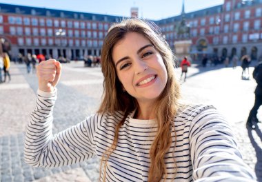 Beautiful young caucasian woman happy and excited in Plaza Mayor Madrid holding the camera and taking a photo of herself. Looking cheerful and joyful. In tourism, European city, travel in Europe. clipart