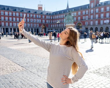 Beautiful young caucasian student tourist woman taking a selfie or video for her tourism and travel around the world blog web in Plaza Mayor Madrid Spain. In social media and vacations in europe. clipart