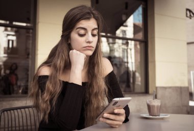 Genç ve güzel kadın şık siyah top ile seyir sıkılmış kahve içme, kahve dükkanında dışında akıllı telefon iletileri gönderme. Yorgun ve donuk duygu. Yaşam tarzı ve teknoloji kavram kullanmak.