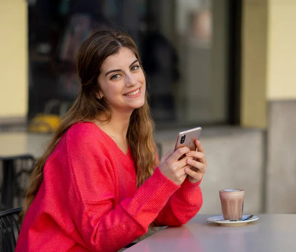 Pretty young fashion woman looking cheerful and happy chatting on social media on smart phone app while drinking coffee in urban city. In lifestyle mobile use and online app and technology concept.