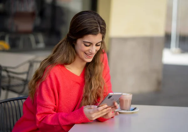 Pretty Happy young woman chatting and using smart mobile phone app looking cheerful drinking coffee in a urban coffee terrace. In online dating, mobile app, internet, connections and technology use.