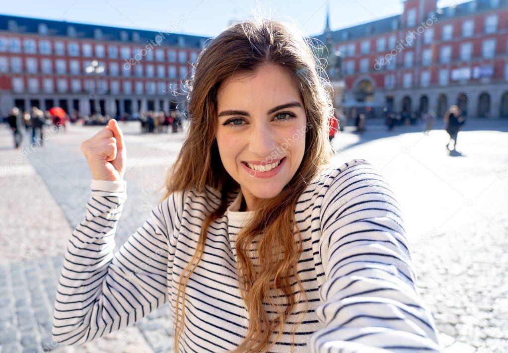 Beautiful young caucasian woman happy and excited in Plaza Mayor Madrid holding the camera and taking a photo of herself. Looking cheerful and joyful. In tourism, European city, travel in Europe.