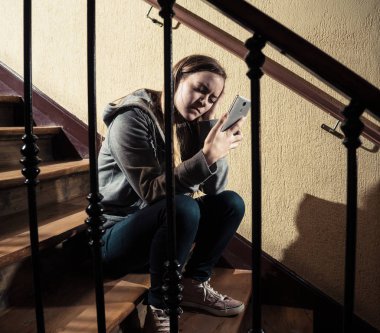 Yalnız, mutsuz, umutsuz ve istismar hissi merdiven üzerinde mobil akıllı telefon siting tarafından siber zorbalık üzücü depresif genç kız kızı kurbanı. Sosyal medya uygulamasında kısa mesaj ile bullied. koyu ışık