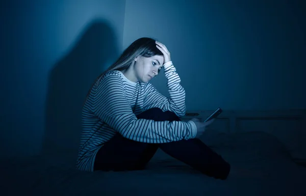 Sad desperate young teenager female girl on smart phone suffering from online bulling and harassment felling lonely and hopeless sitting on bed at night. CYberbullying and dangers of internet concept.