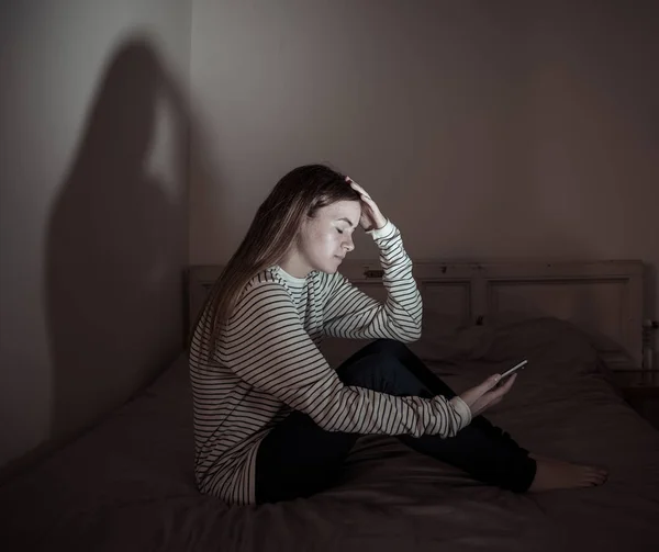 モバイル携帯電話中毒に苦しむ若いティーンエイジャーの女の子夜遅くにベッドに座っていることを必要とする不眠症を感じて寂しいと落ち込んでいます インチ — ストック写真