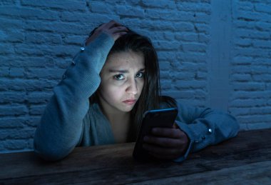 Dramatic portrait of sad scared young woman victim of online harassment and cyberbullying. looking at smart mobile phone stressed and in fear being online abused by stalker. In Dangers of internet. clipart