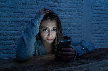 Dramatic portrait of sad scared young woman victim of online harassment and cyberbullying. looking at smart mobile phone stressed and in fear being online abused by stalker. In Dangers of internet. clipart