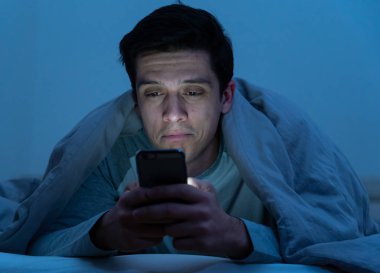 Genç cep telefonu bağımlısı adam uyanık gece yatakta sohbet için akıllı telefon kullanarak, flört ve metin mesajı gönderme. İletişim, Internet bağımlılığı ve sosyal medya ağ istismarı kavramı