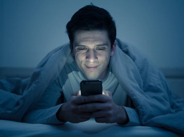 Genç cep telefonu bağımlısı adam uyanık gece yatakta sohbet için akıllı telefon kullanarak, flört ve metin mesajı gönderme. İletişim, Internet bağımlılığı ve sosyal medya ağ istismarı kavramı