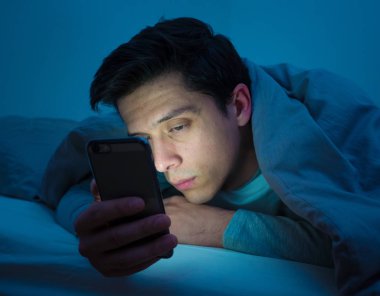Genç cep telefonu bağımlısı adam uyanık gece yatakta sohbet için akıllı telefon kullanarak, flört ve metin mesajı gönderme. İletişim, Internet bağımlılığı ve sosyal medya ağ istismarı kavramı