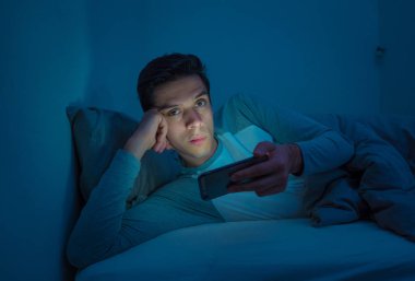 Genç cep telefonu bağımlısı adam uyanık gece yatakta sohbet için akıllı telefon kullanarak, flört ve metin mesajı gönderme. İletişim, Internet bağımlılığı ve sosyal medya ağ istismarı kavramı
