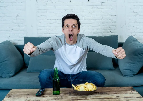 Lifestyle portrait of excited football fan having fun watching soccer or football game on television. Enjoying and celebrating goal and victory drinking beer and eating chips. Sports fans and Goal.