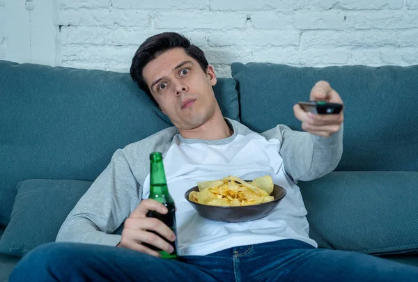 Lifestyle portrait of young bored man on couch with remote control zapping for movie or live sport. Looking disinterested drinking beer. Sedentary and mass social media or Television addiction.