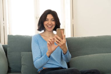 Mutlu genç kadın evde kanepede oturan onu akıllı telefon kullanarak. Boş zaman ve mobil bağımlılık