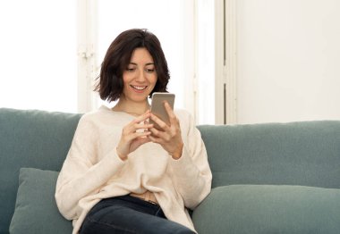 Mutlu genç kadın evde kanepede oturan onu akıllı telefon kullanarak. Boş zaman ve mobil bağımlılık
