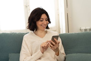 Mutlu genç kadın evde kanepede oturan onu akıllı telefon kullanarak. Boş zaman ve mobil bağımlılık