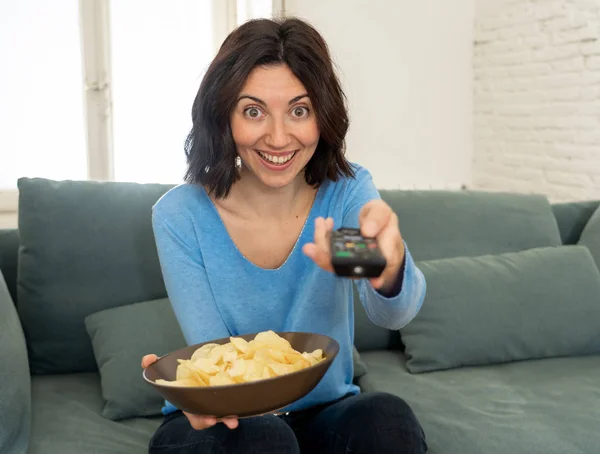 Lifestyle portrait of cheerful young woman sitting on the couch watching TV holding remote control