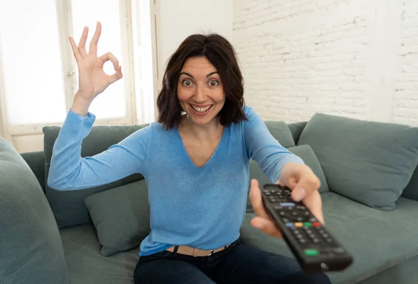 Lifestyle portrait of cheerful young woman sitting on the couch watching TV holding remote control — Stock Photo, Image