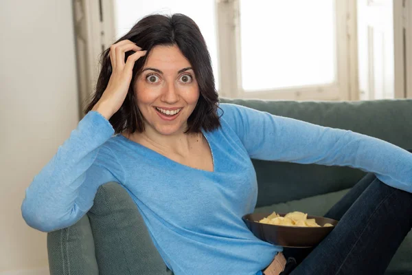 Lifestyle portrait of cheerful young woman sitting on the couch watching TV holding remote control — Stock Photo, Image