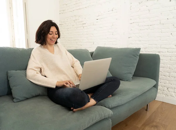 Eine fröhliche junge Frau sitzt im Schneidersitz auf dem Sofa und arbeitet an ihrem Laptop. im Technologiekonzept — Stockfoto