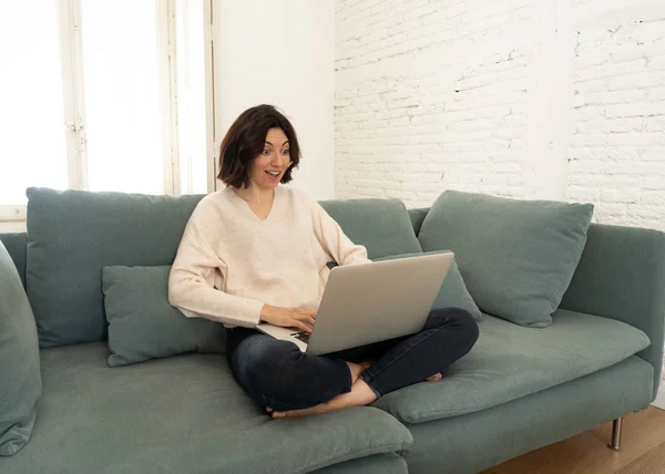 Щаслива молода жінка працює на ноутбуці, сидячи схрещеними ногами на дивані. У концепції технології — стокове фото