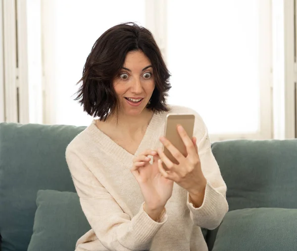 Happy young woman using her smart phone sitting on sofa at home. In leisure and mobile addiction — Stock Photo, Image