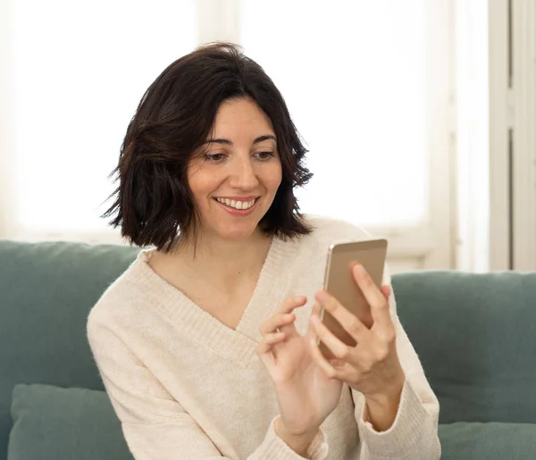 Счастливая молодая женщина использует свой смартфон, сидя дома на диване. В досуге и мобильной зависимости — стоковое фото