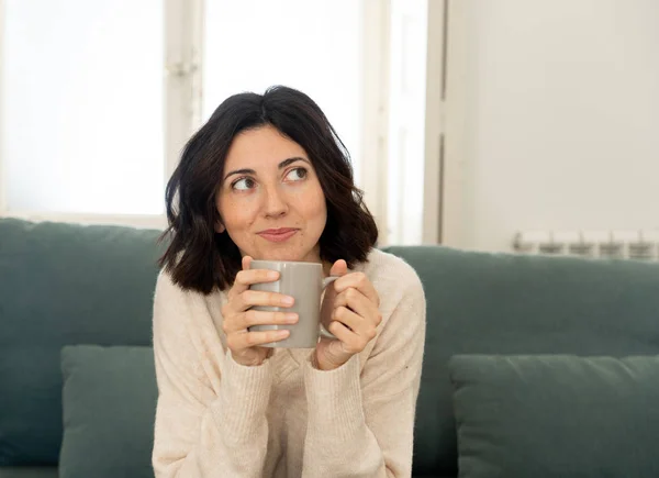 自宅のソファに熱い飲み物を持って座っている幸せな若い女性。余暇と空き時間のコンセプトで — ストック写真